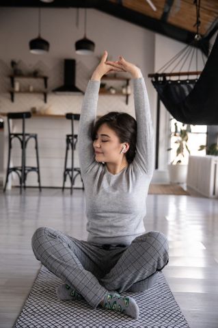Cheerful young ethnic female in earbuds sitting on yoga mat with crossed legs and closed eyes while listening to music and raising hands in apartment