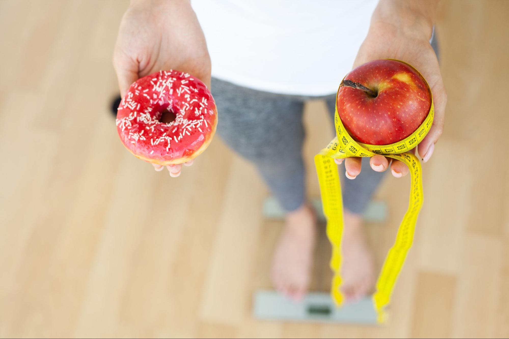 person holding donut and apple with measuring tape while standing on a weighing scale 