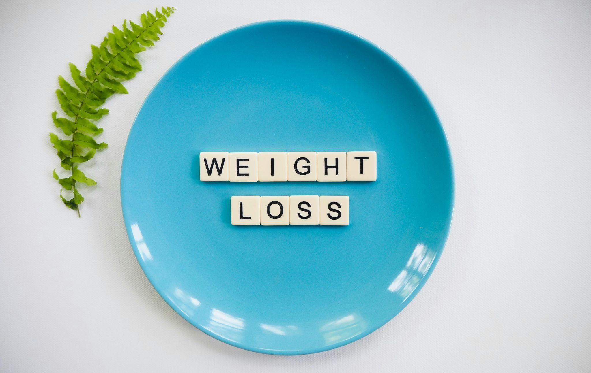 A blue plate with white letter tiles spelling "WEIGHT LOSS" placed in the center. The plate is set on a white surface, and a green fern leaf lies to the left of the plate.