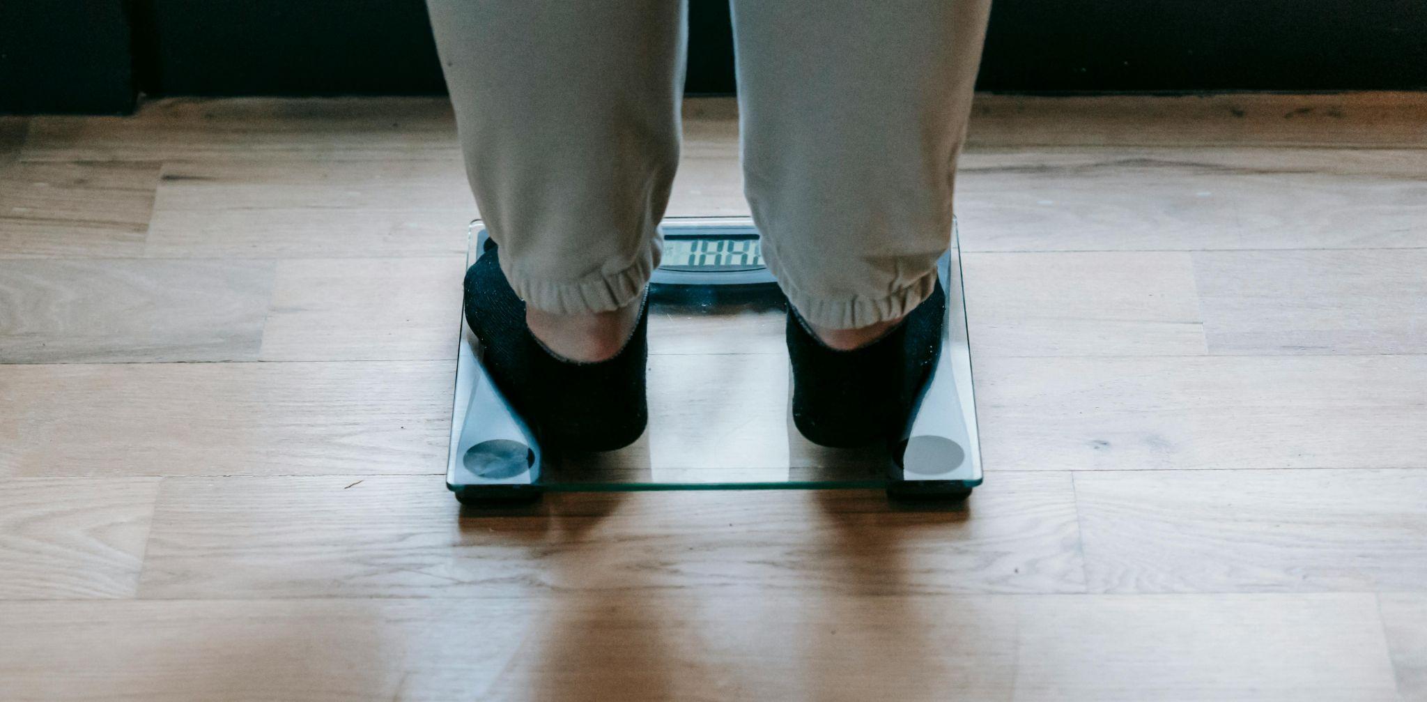 Person standing on a digital glass weighing scale, wearing black socks and beige pants, on a wooden floor. The display on the scale is partially visible.