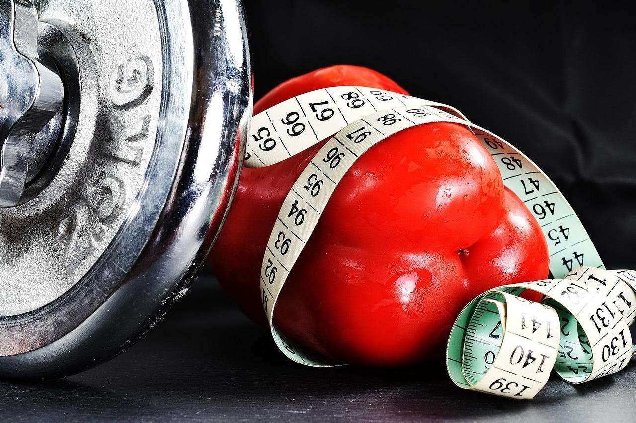 A shiny silver dumbbell plate is on the left, and a bright red bell pepper wrapped with a white measuring tape is on the right, all set against a dark background.