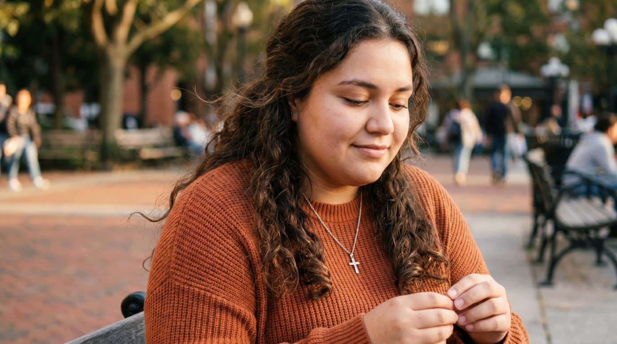 Close-up shot of a young woman with curly hair wearing an orange sweater and cross necklace, sitting outside on a park bench looking down thoughtfully, illustrating a patient preparing for or reflecting on treatment.