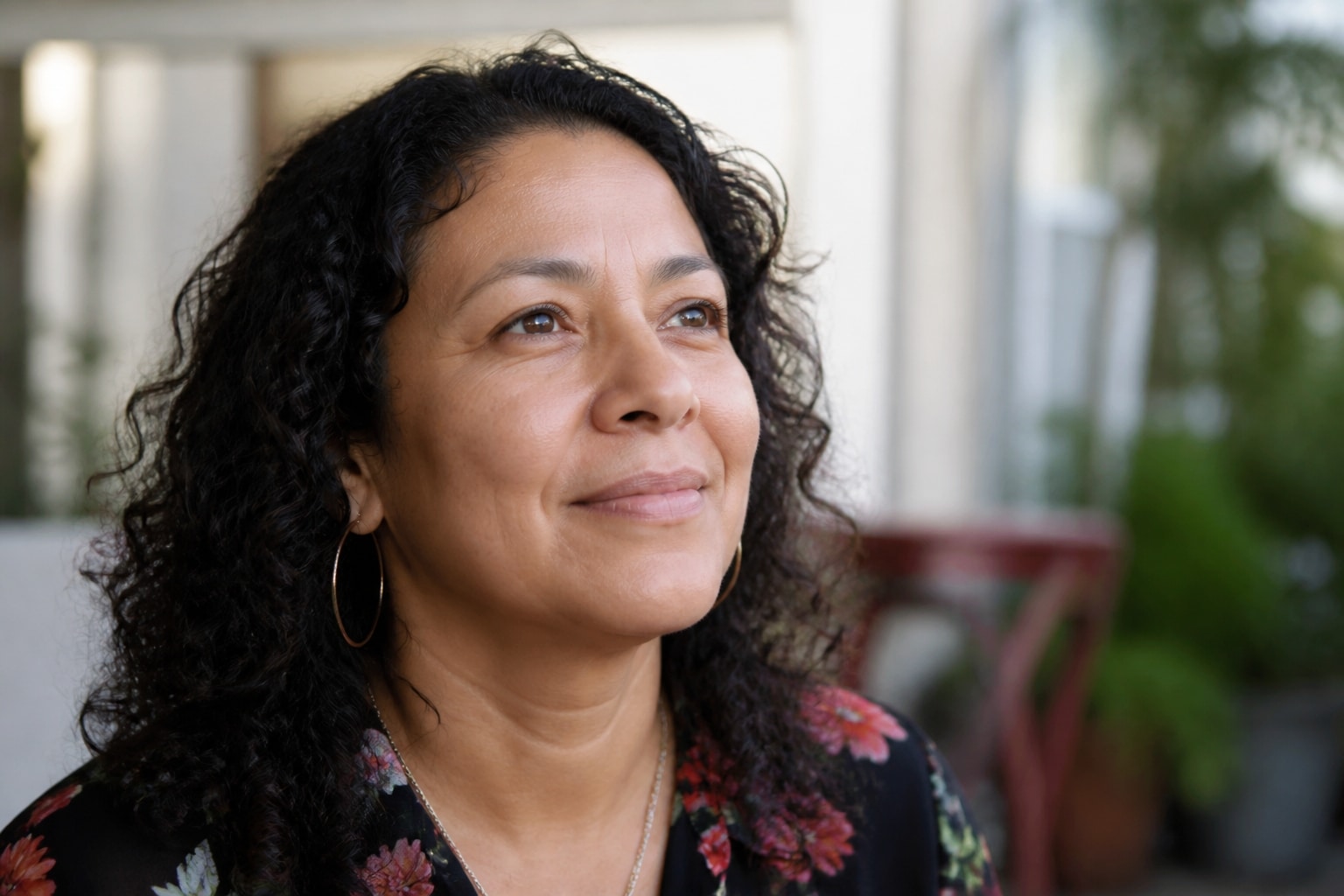 A middle-aged Hispanic woman with dark curly hair and hoop earrings, smiling with gentle optimism while looking slightly off-camera.